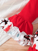 Little girl wearing a red long-sleeve Christmas dress with reindeer print, layered ruffle skirt and festive patterned sleeve cuffs beside a decorated Christmas tree.