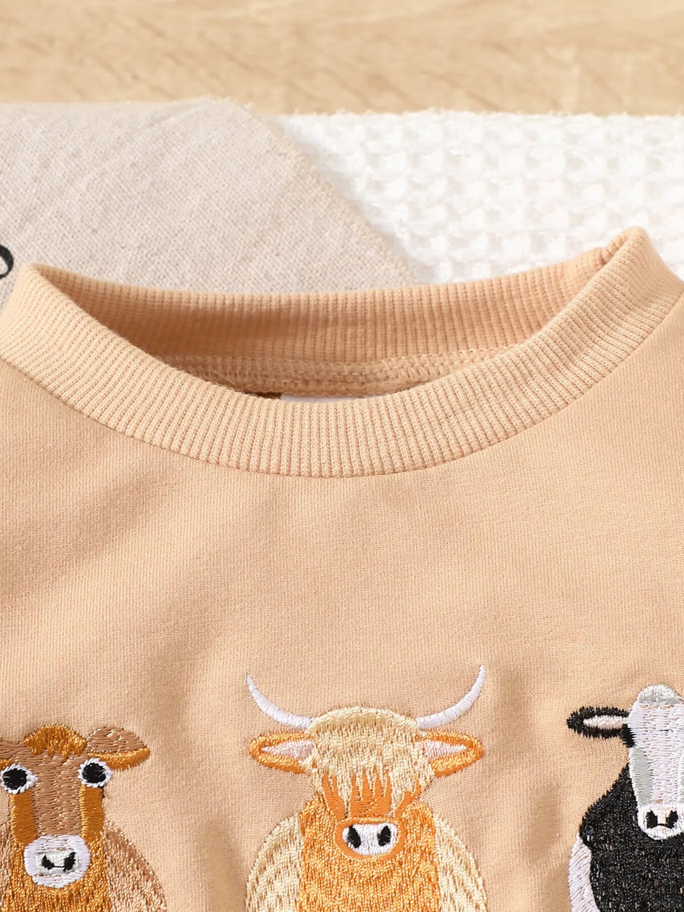 Children's two-piece farm animal outfit neckline featuring a beige long-sleeve sweatshirt with embroidered cows and coordinating brown and cream cow-print drawstring pants.