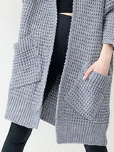 Women's gray chunky waffle knit open front cardigan with front pockets, cozy oversized fall sweater layered over a black top and leggings.