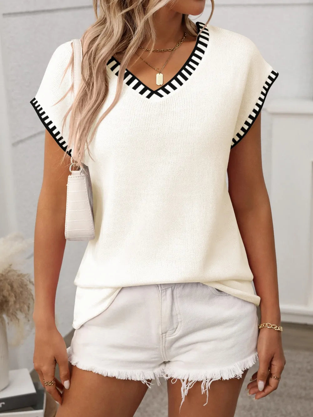 Woman wearing a cream short sleeve knit top with black contrast trim along the V-neckline and sleeves.