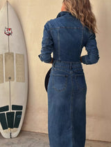 Woman wearing a dark blue button-front denim midi dress with gold-tone buttons, collared neckline, patch pockets, contrast stitching and front slit.