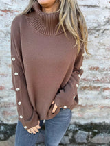 Woman wearing a dark brown ribbed turtleneck sweater with decorative button sleeves and jeans.