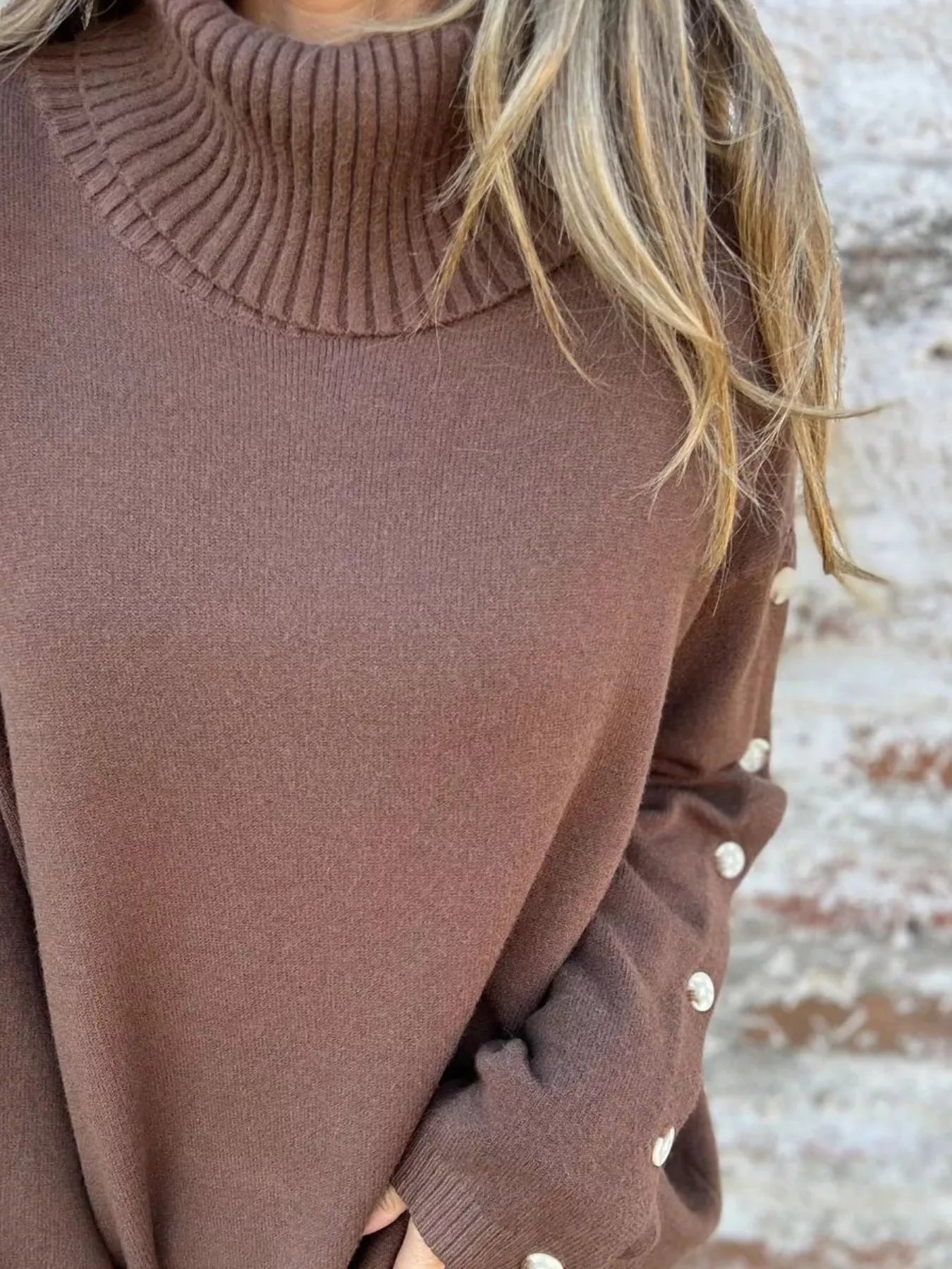Woman wearing a dark brown ribbed turtleneck sweater with decorative button sleeves and jeans.