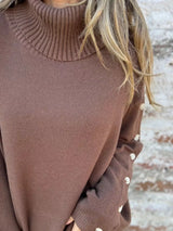 Woman wearing a dark brown ribbed turtleneck sweater with decorative button sleeves and jeans.