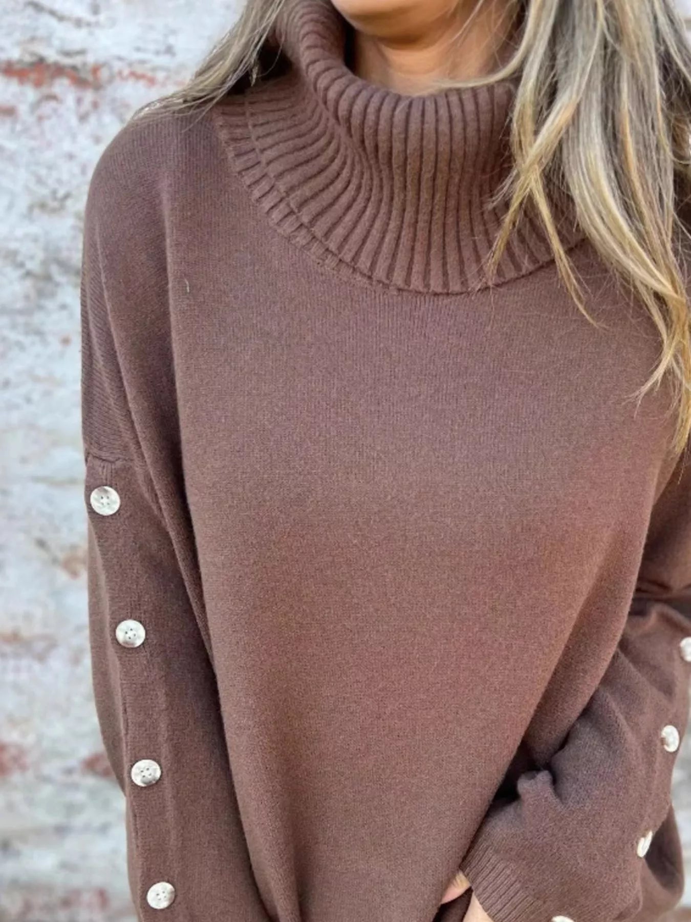 Woman wearing a dark brown ribbed turtleneck sweater with decorative button sleeves and jeans.