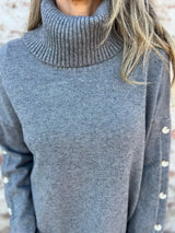 Woman wearing a dark gray ribbed turtleneck sweater with decorative button sleeves and jeans.