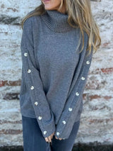 Woman wearing a dark gray ribbed turtleneck sweater with decorative button sleeves and jeans.
