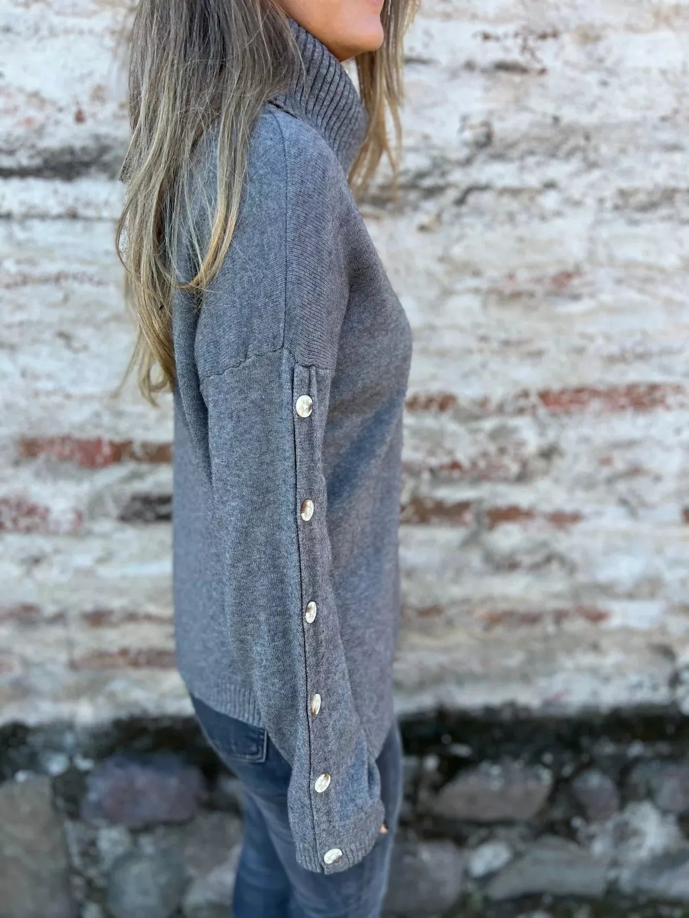 Woman wearing a dark gray ribbed turtleneck sweater with decorative button sleeves and jeans.