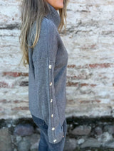 Woman wearing a dark gray ribbed turtleneck sweater with decorative button sleeves and jeans.