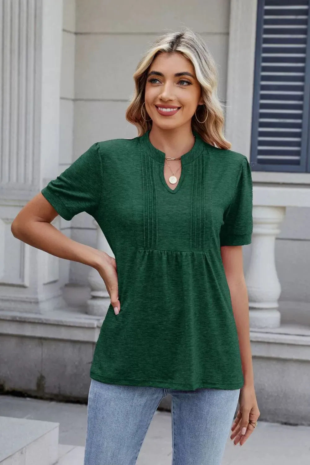 Woman wearing a dark green notched short-sleeve top with light-wash jeans, sunglasses, and layered jewelry.