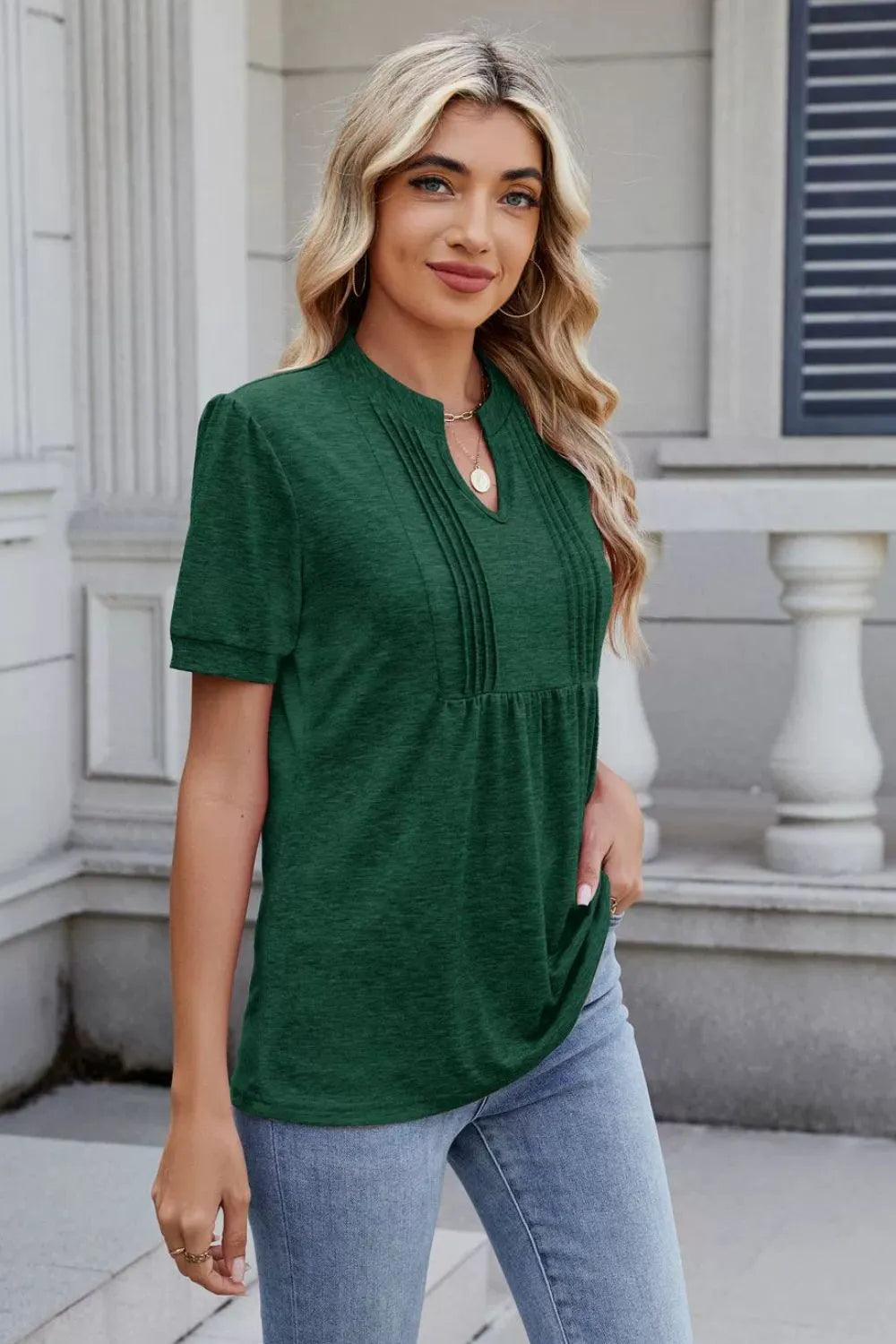 Woman wearing a dark green notched short-sleeve top with light-wash jeans, sunglasses, and layered jewelry.