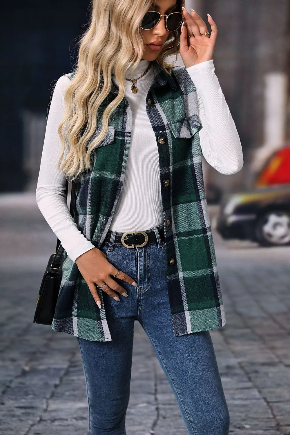 Woman wearing a dark green plaid button-up vest coat layered over a white turtleneck with blue jeans and gold jewelry for a cozy fall outfit.