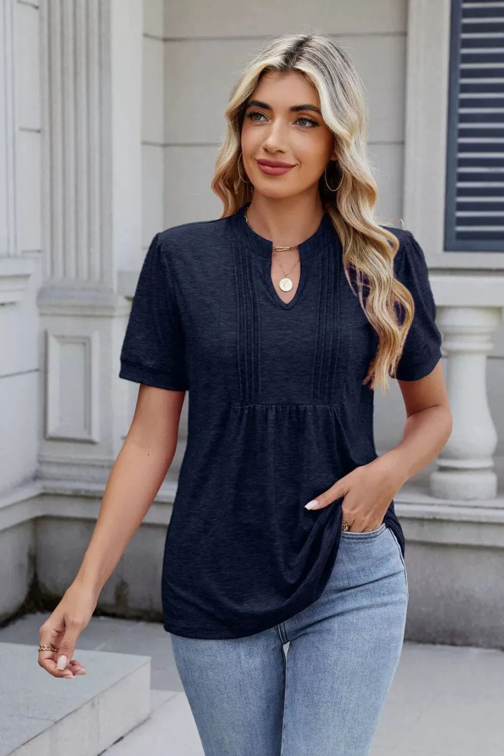 Woman wearing a dark navy notched short-sleeve top with light-wash jeans, sunglasses, and layered jewelry.