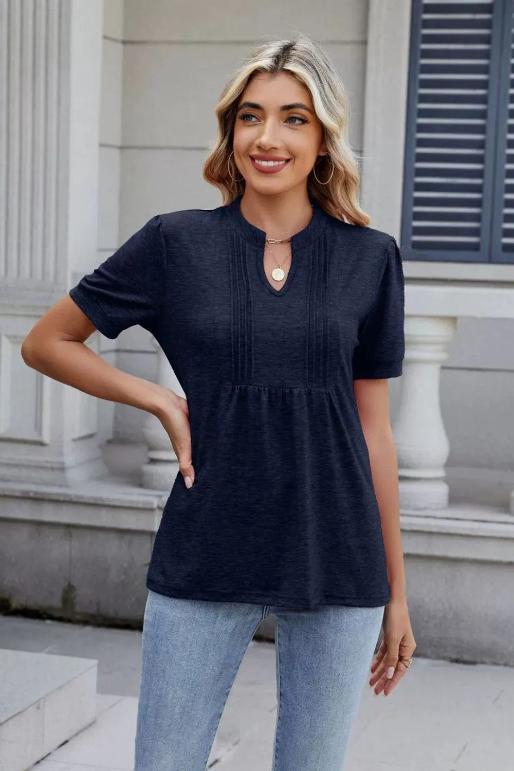 Woman wearing a dark navy notched short-sleeve top with light-wash jeans, sunglasses, and layered jewelry.