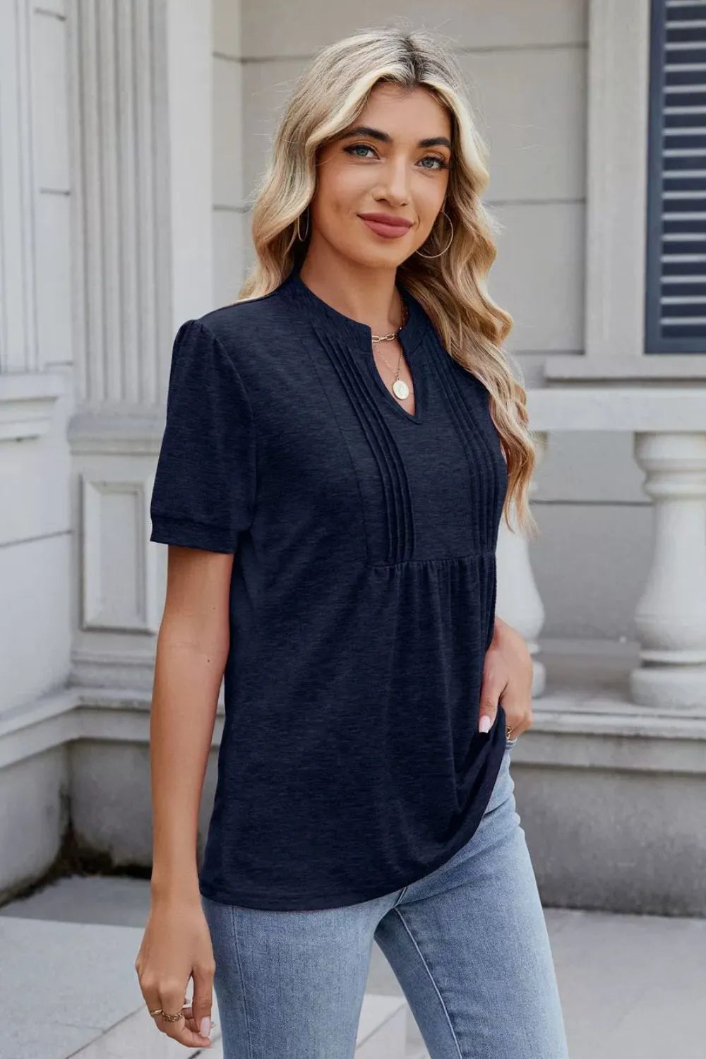 Woman wearing a dark navy notched short-sleeve top with light-wash jeans, sunglasses, and layered jewelry.