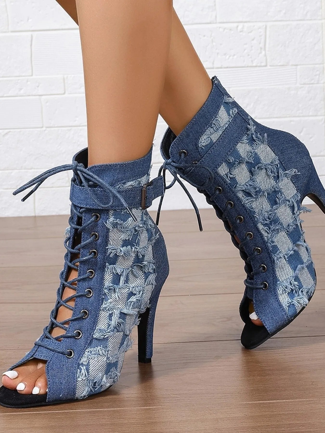 Blue denim lace-up high-heeled boots on a wooden floor with a white brick wall background.
