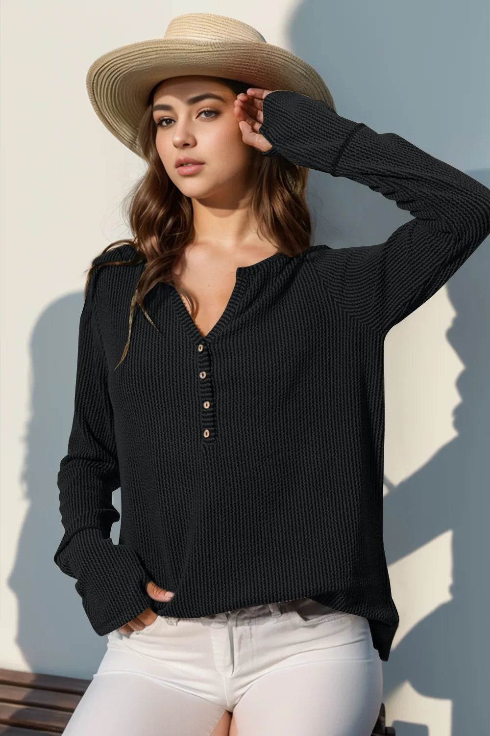 Woman wearing a black ribbed sweater and white pants with a straw hat against a light background