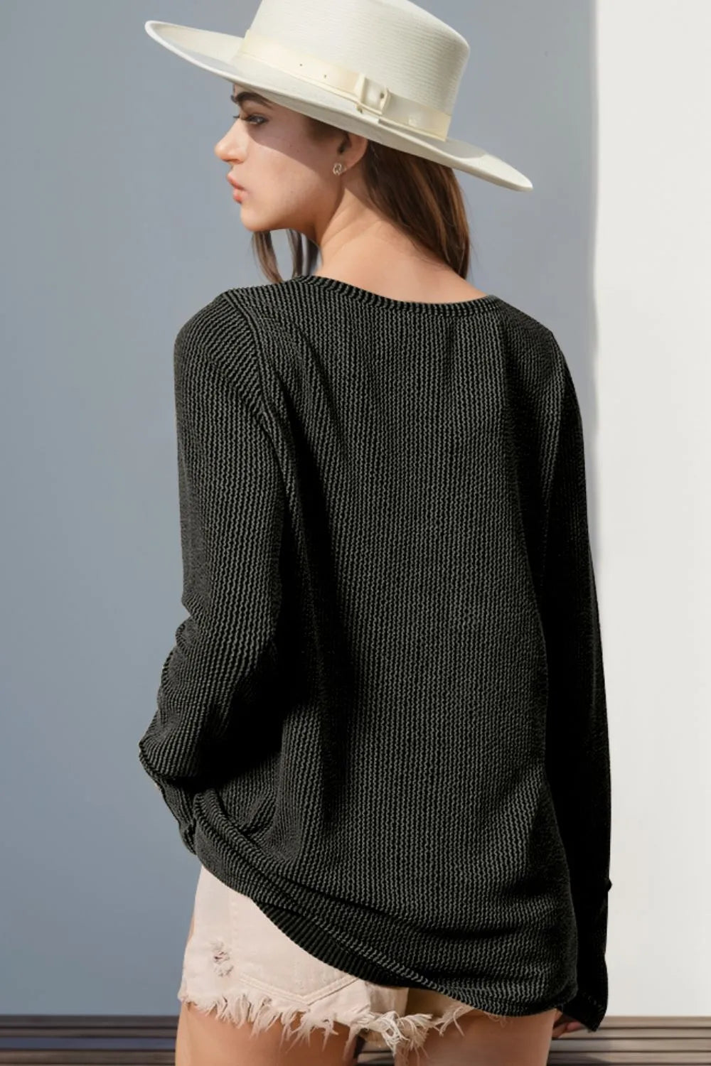 Woman wearing a dark gray textured sweater and white hat against a gray wall.