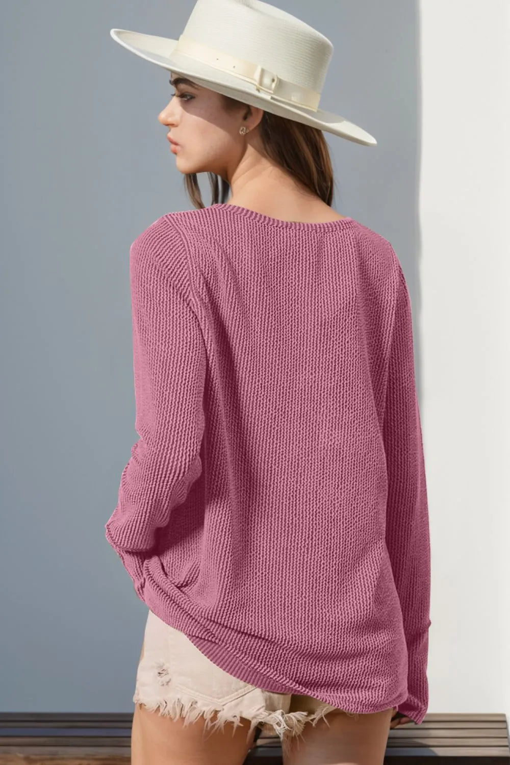 Woman wearing a pink sweater and white hat against a gray wall.