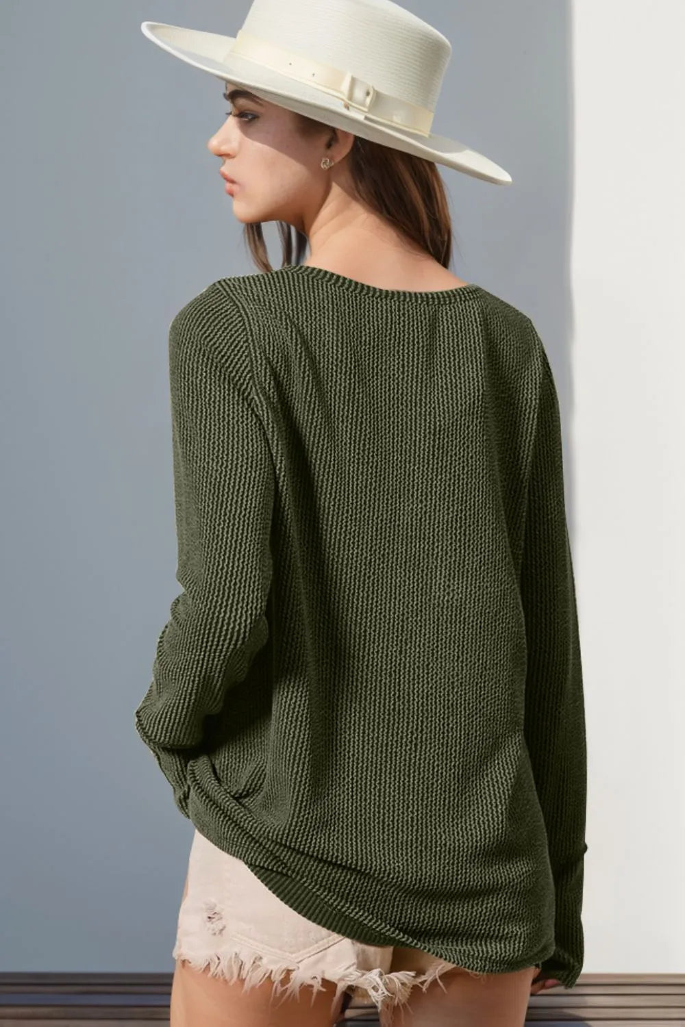 Person wearing a green sweater and white hat against a gray wall showing back of shirt.