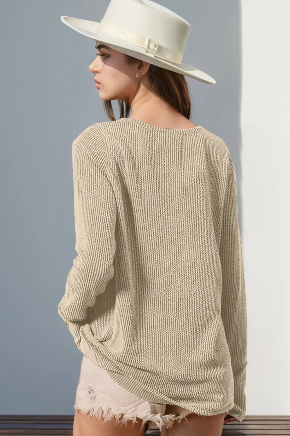 Woman wearing a beige knitted sweater and white hat against a gray wall.