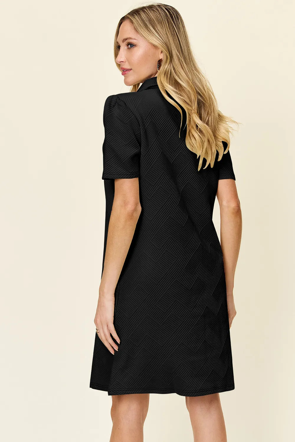 Woman wearing a black dress against a beige background showing back of the dress.