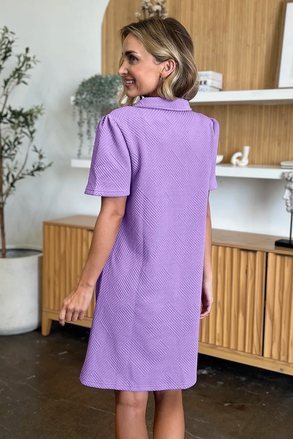 Woman wearing a purple dress in an indoor setting with wooden cabinets and plants showing the back of the dress.