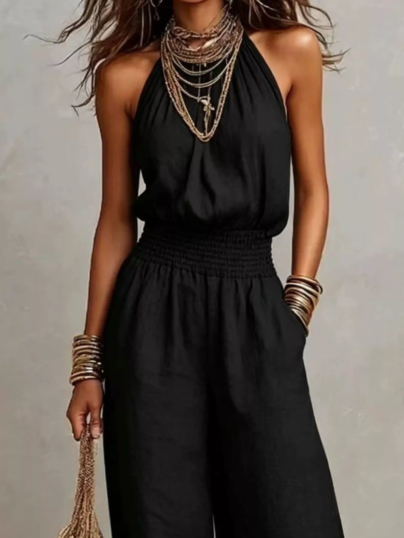 Woman wearing a black halter-neck wide-leg jumpsuit with a smocked waist, layered gold jewelry, and bracelets, styled for an elegant evening look.