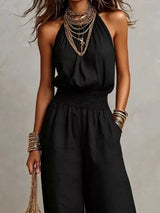 Woman wearing a black halter-neck wide-leg jumpsuit with a smocked waist, layered gold jewelry, and bracelets, styled for an elegant evening look.