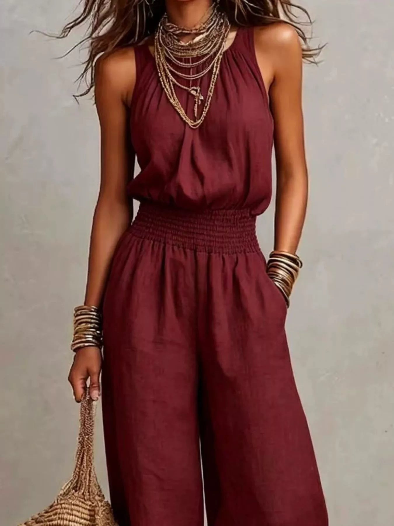 Woman wearing a burgundy halter-neck wide-leg jumpsuit with a smocked waist, layered gold jewelry, and bracelets, styled for an elegant evening look.
