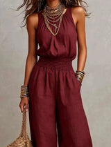 Woman wearing a burgundy halter-neck wide-leg jumpsuit with a smocked waist, layered gold jewelry, and bracelets, styled for an elegant evening look.