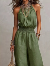 Woman wearing a green halter-neck wide-leg jumpsuit with a smocked waist, layered gold jewelry, and bracelets, styled for an elegant evening look.