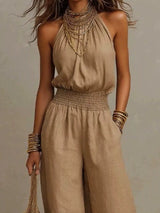Woman wearing a light brown halter-neck wide-leg jumpsuit with a smocked waist, layered gold jewelry, and bracelets, styled for an elegant evening look.