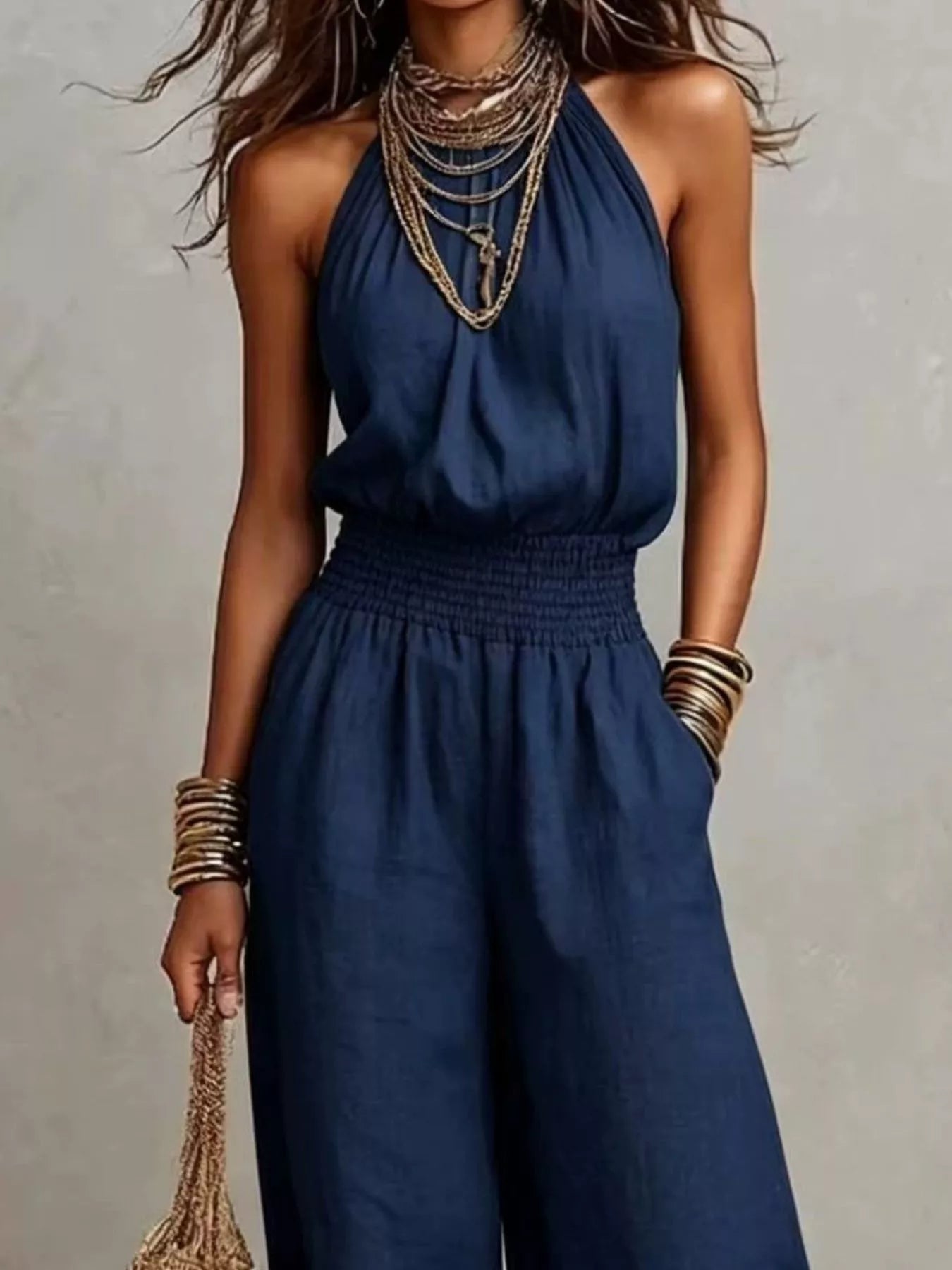 Woman wearing a navy-blue halter-neck wide-leg jumpsuit with a smocked waist, layered gold jewelry, and bracelets, styled for an elegant evening look.