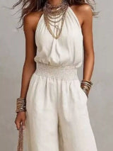 Woman wearing a white halter-neck wide-leg jumpsuit with a smocked waist, layered gold jewelry, and bracelets, styled for an elegant evening look.