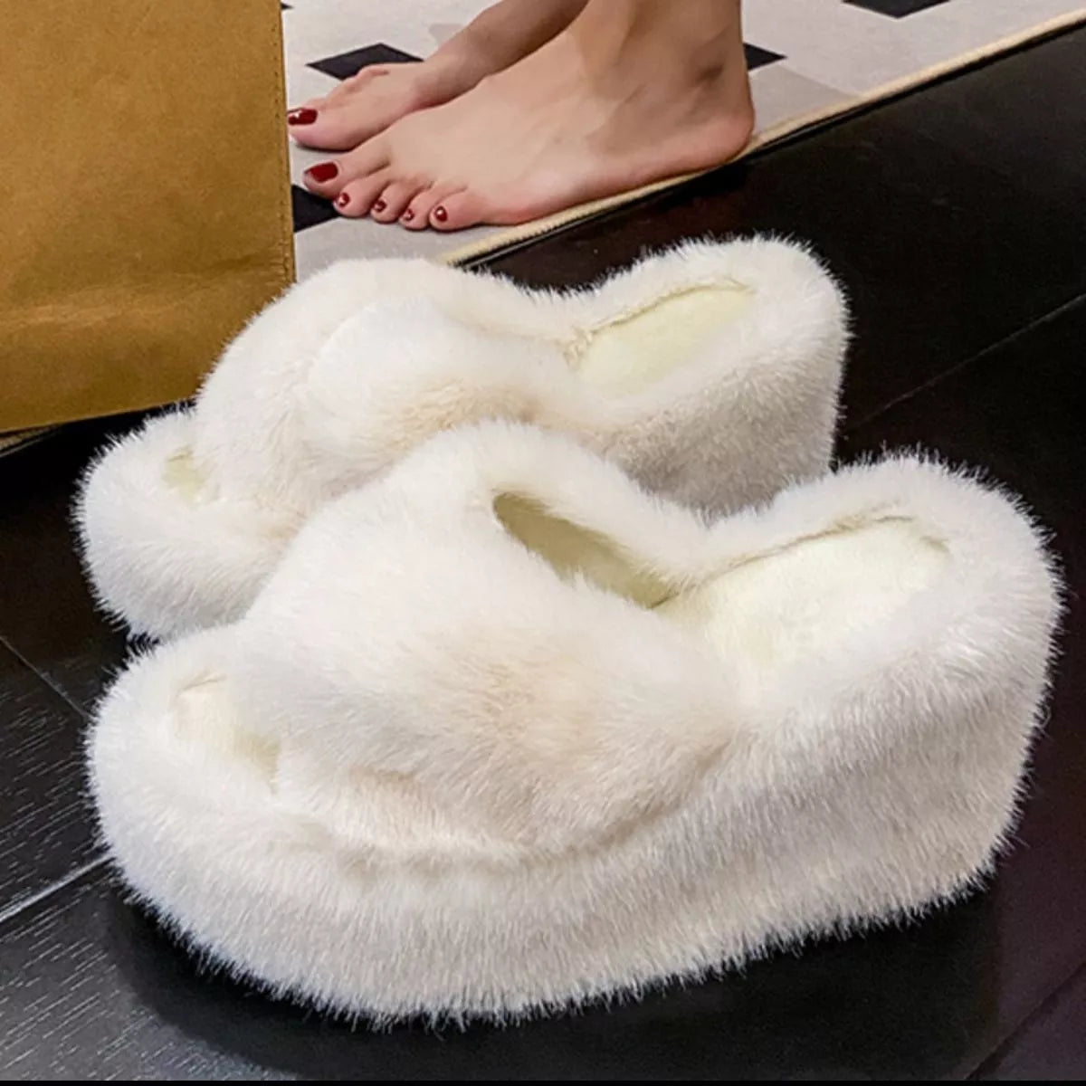 White fuzzy slippers on a dark surface with a person's feet in the background.