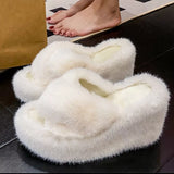 White fuzzy slippers on a dark surface with a person's feet in the background.