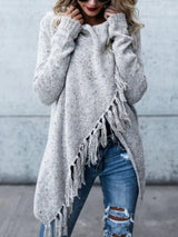Woman wearing a light gray fringe long sleeve wrap cardigan with an asymmetrical draped front over a white top and jeans.