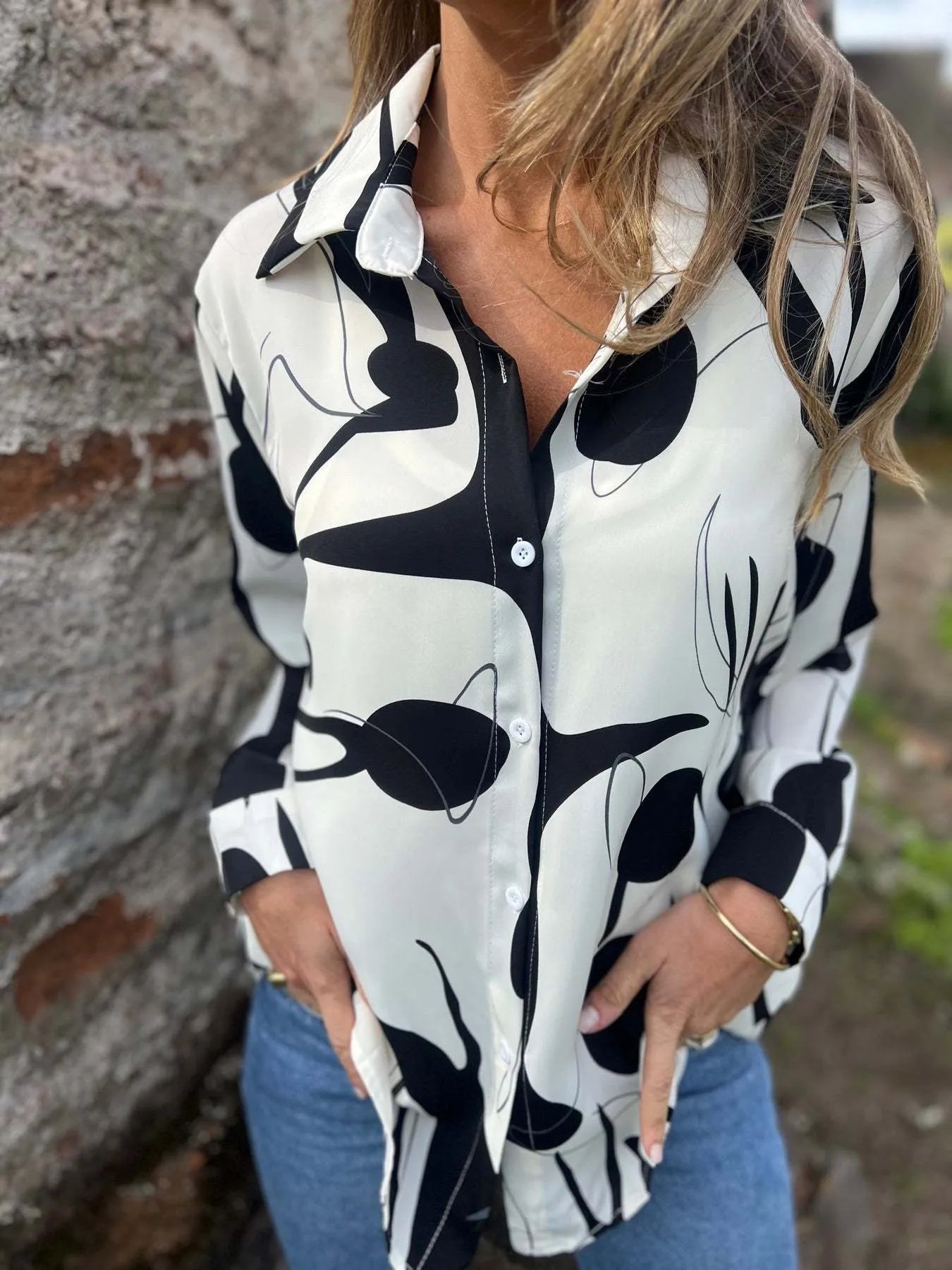 Person wearing a black and white patterned shirt with jeans against a stone wall.