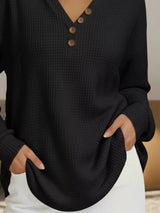 Black textured long-sleeve top worn by a person with hands in pockets, blurred background