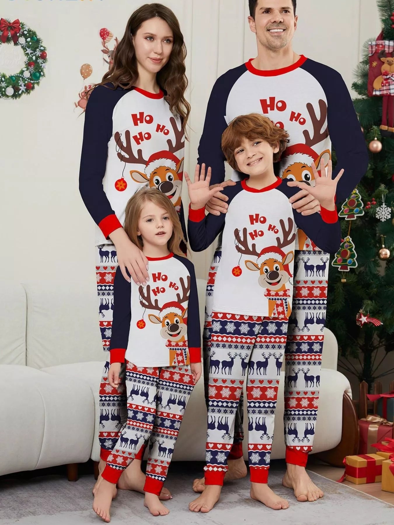 Family of four wearing matching Christmas reindeer pajamas with navy sleeves, red trim, smiling Santa-hat reindeer graphics and festive patterned pants.