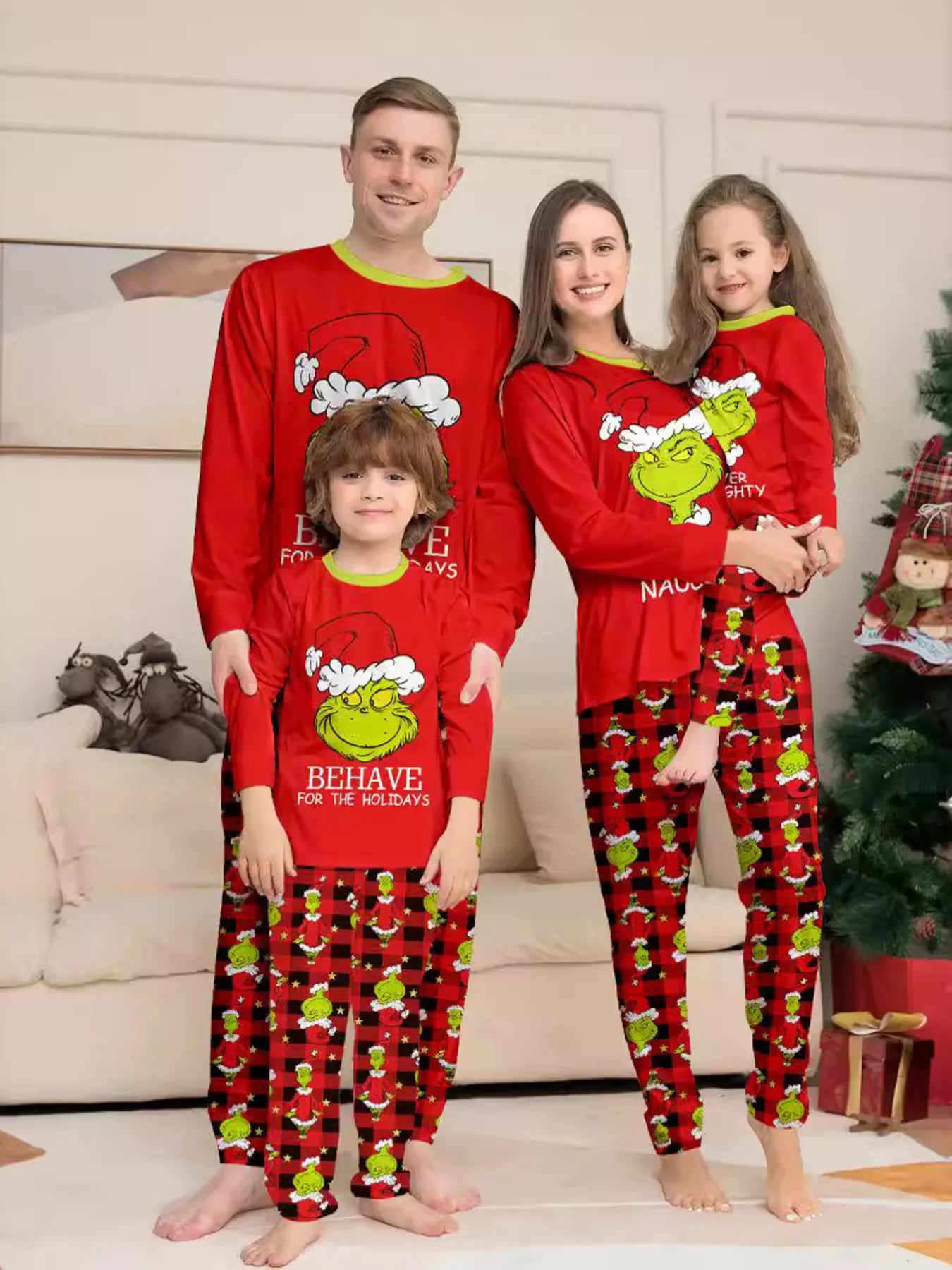 Family of four wearing matching red Grinch-themed Christmas pajama tops and red and black plaid printed pants beside a Christmas tree.