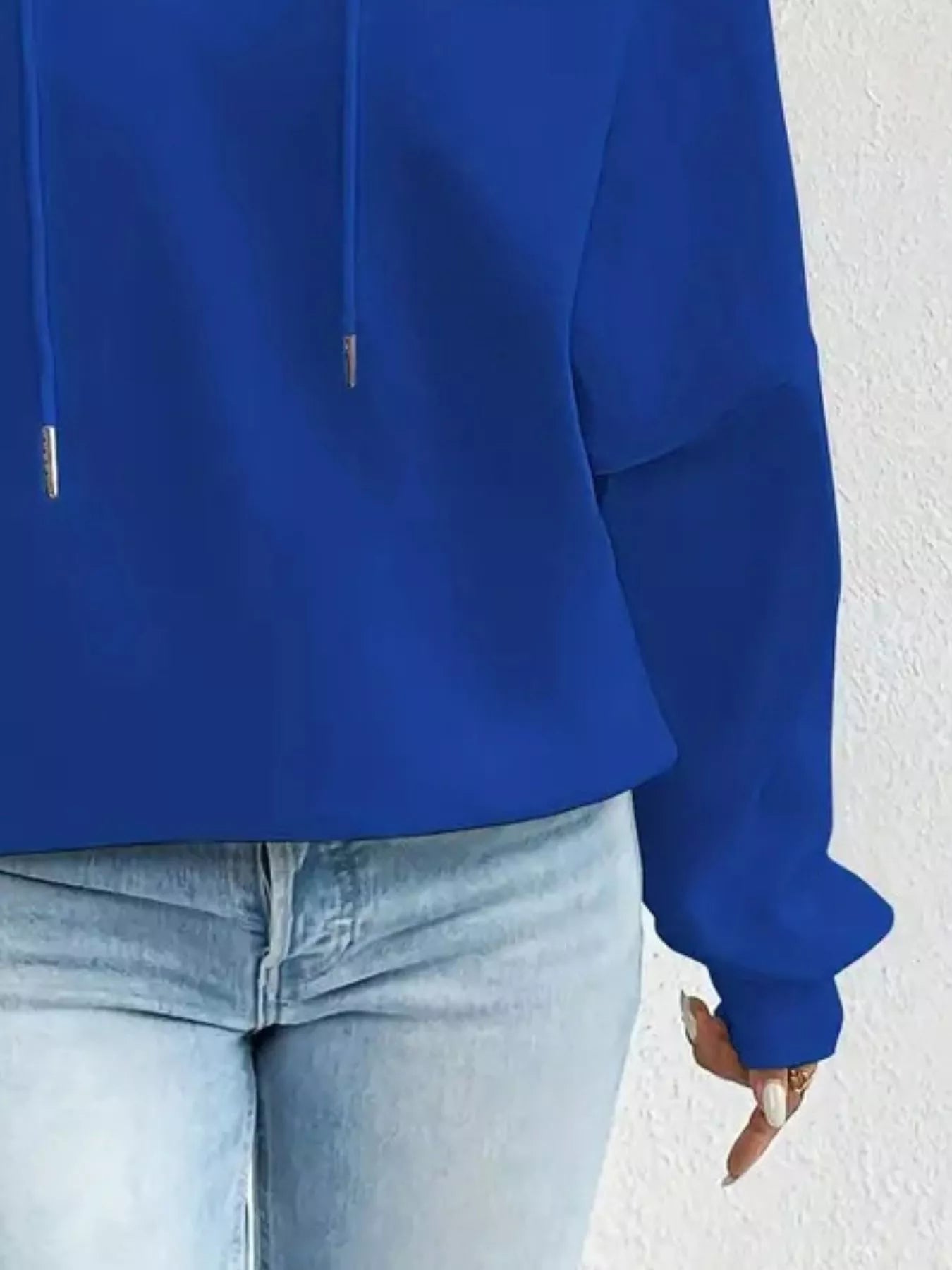 Person wearing a blue hoodie and light blue jeans against a white wall