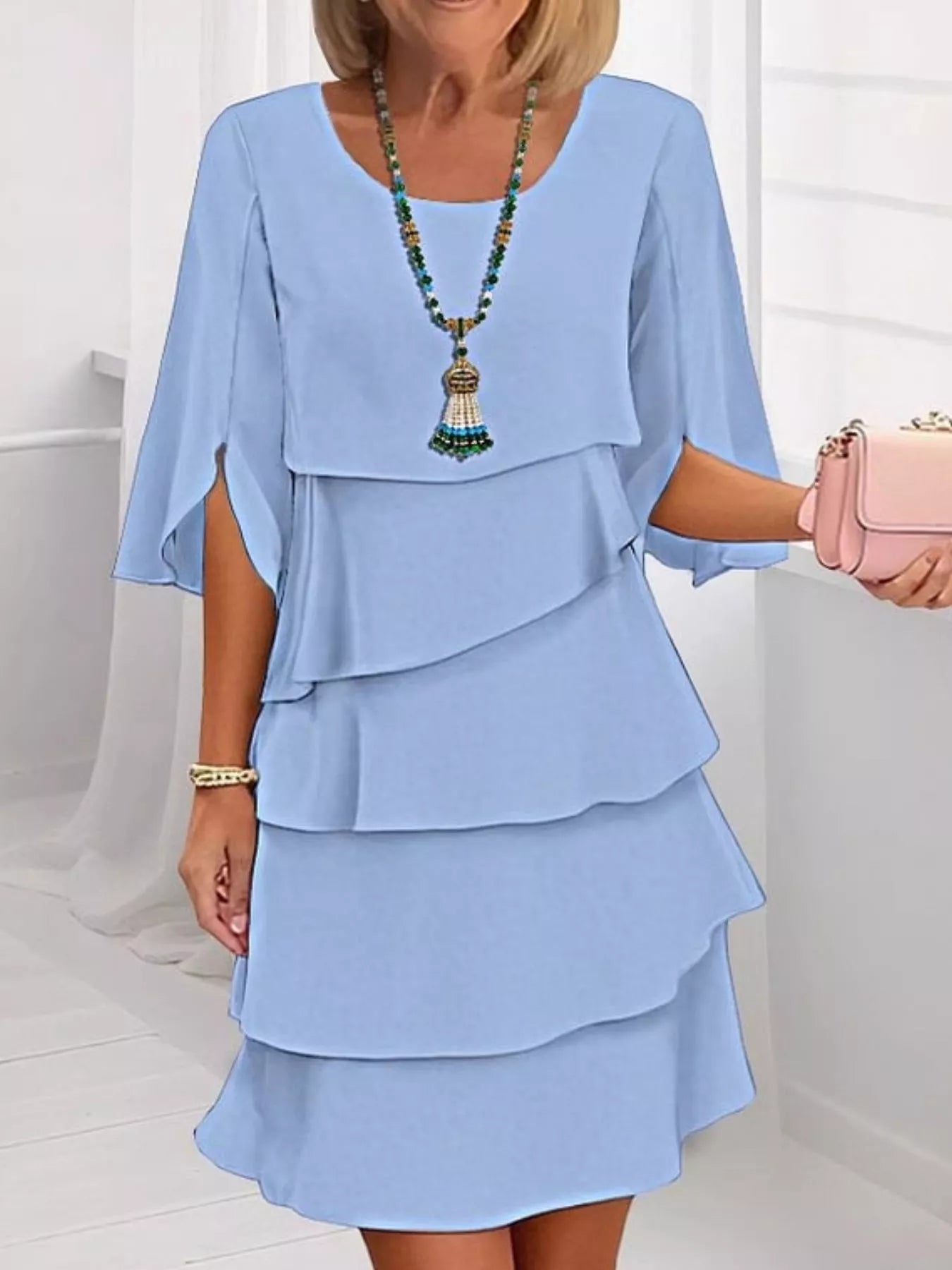 Woman wearing a light blue dress with ruffled layers, holding a pink handbag indoors.