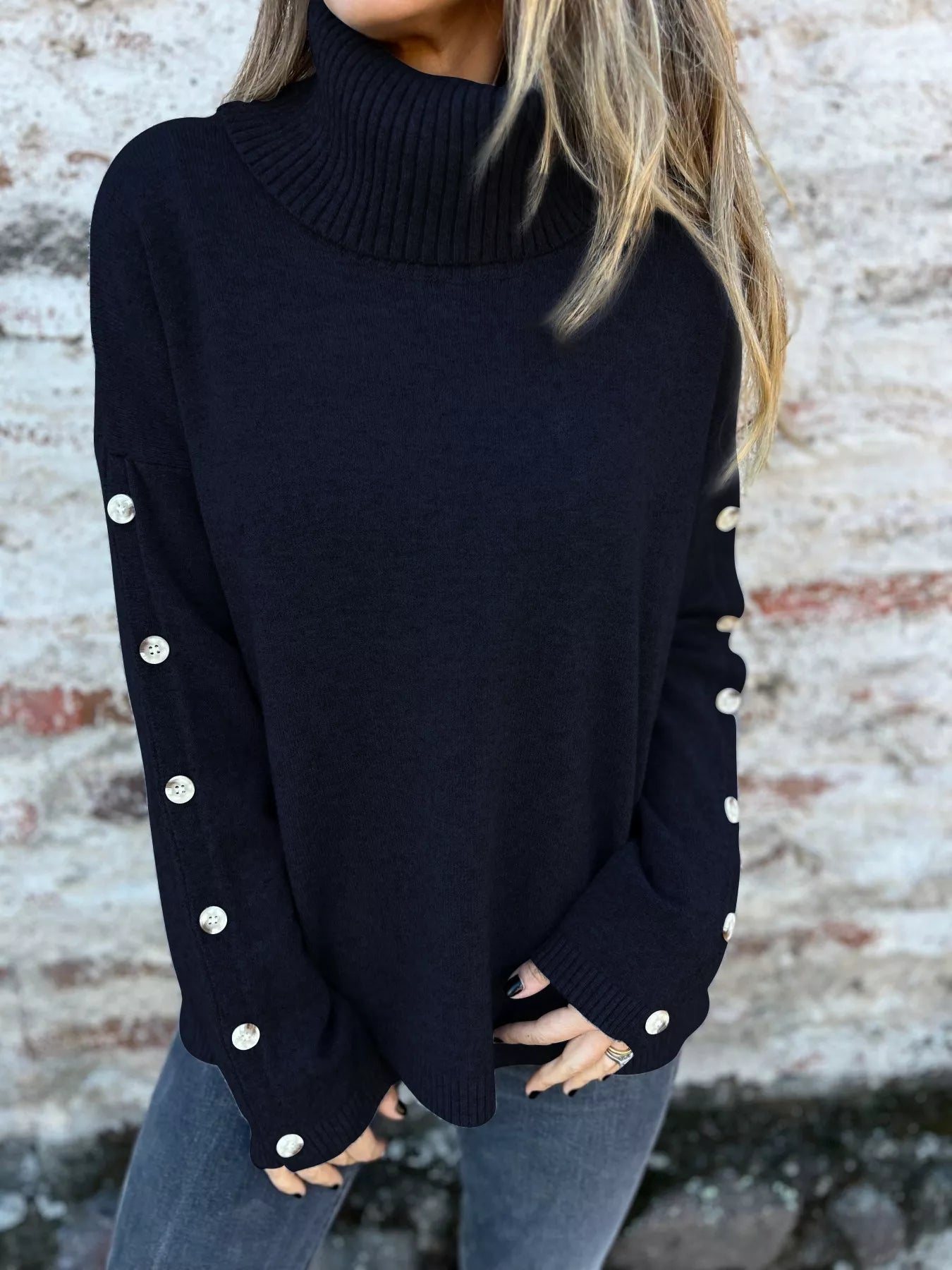 Woman wearing a black ribbed turtleneck sweater with decorative button sleeves and jeans.