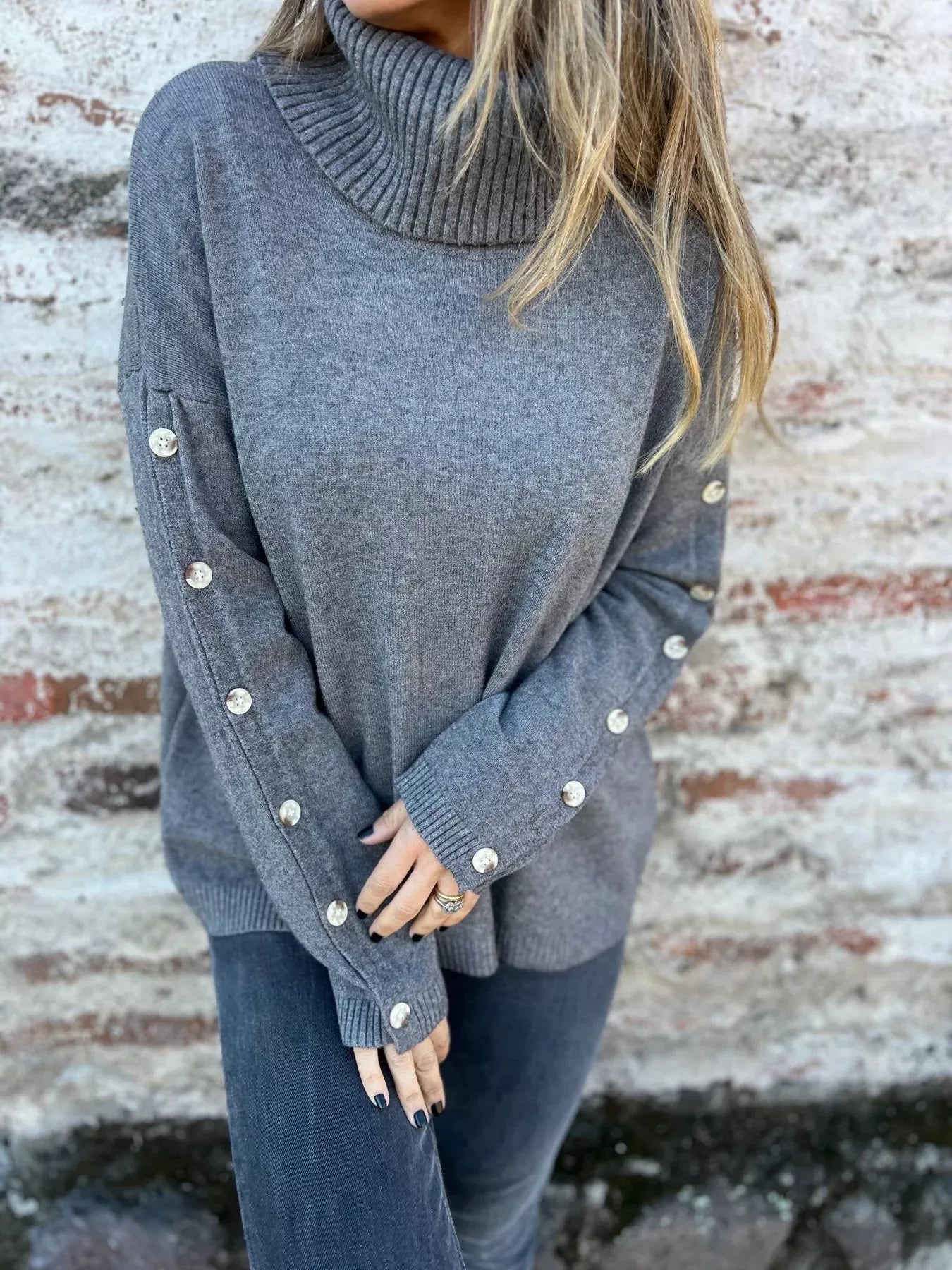 Woman wearing a dark gray ribbed turtleneck sweater with decorative button sleeves and jeans.