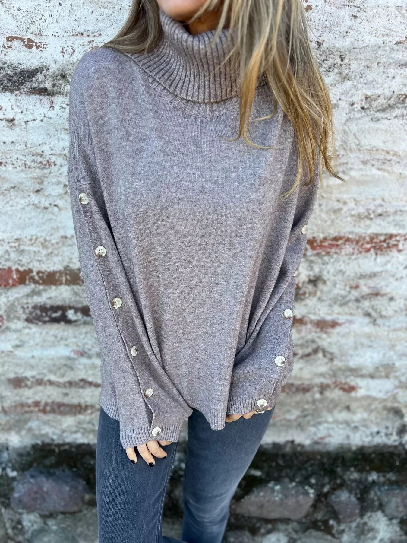 Woman wearing a gray ribbed turtleneck sweater with decorative button sleeves and jeans.