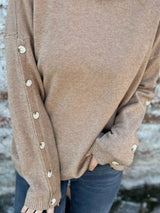 Woman wearing a khaki ribbed turtleneck sweater with decorative button sleeves and jeans.