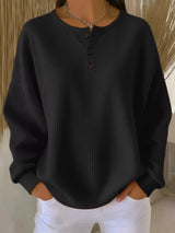 Person wearing a black textured sweater with buttons and white pants against a neutral background