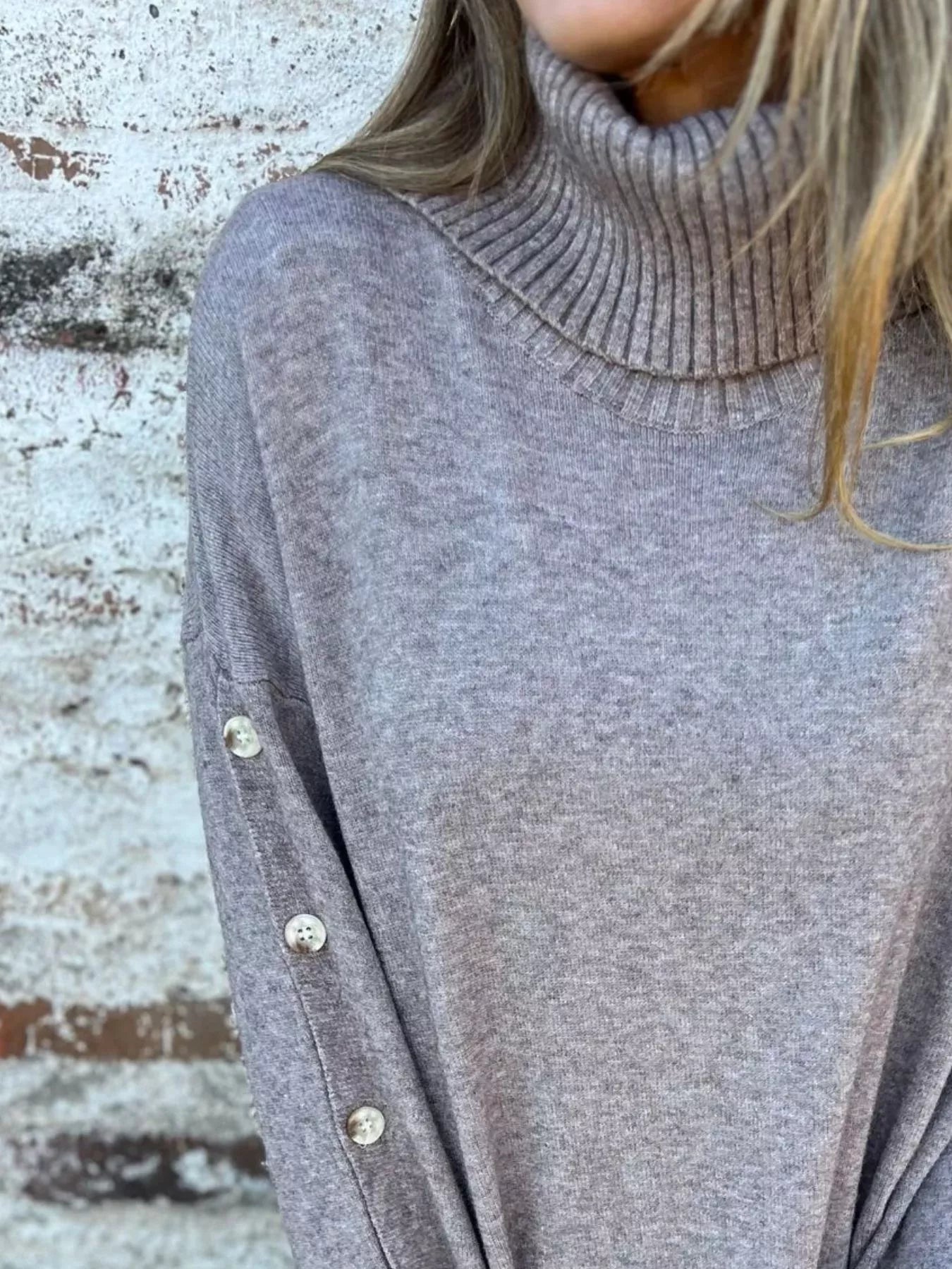 Woman wearing a gray ribbed turtleneck sweater with decorative button sleeves and jeans.